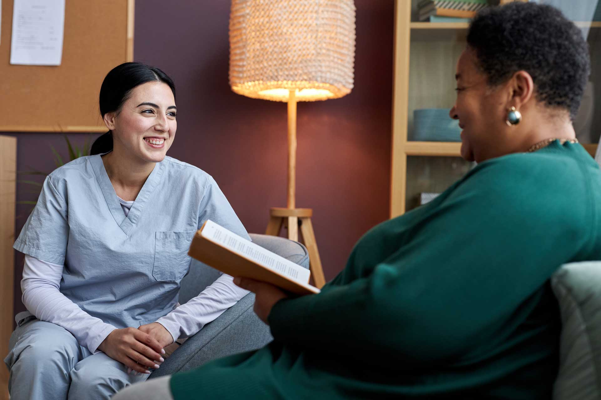 Community_578985590_web two women smiling at each other, one in scrubs and the other reading a book